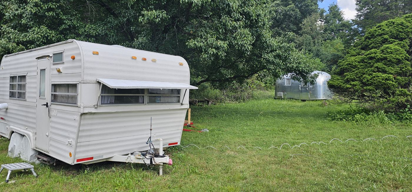 The white vintage camper in the foreground with the Airstream across the clearing