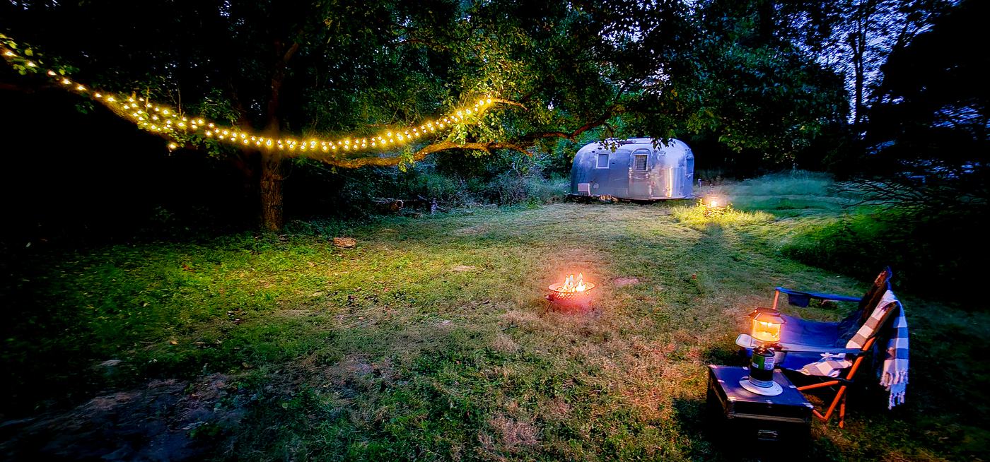 String lights in a big tree at dusk, a small fire, and the Airstream in the background