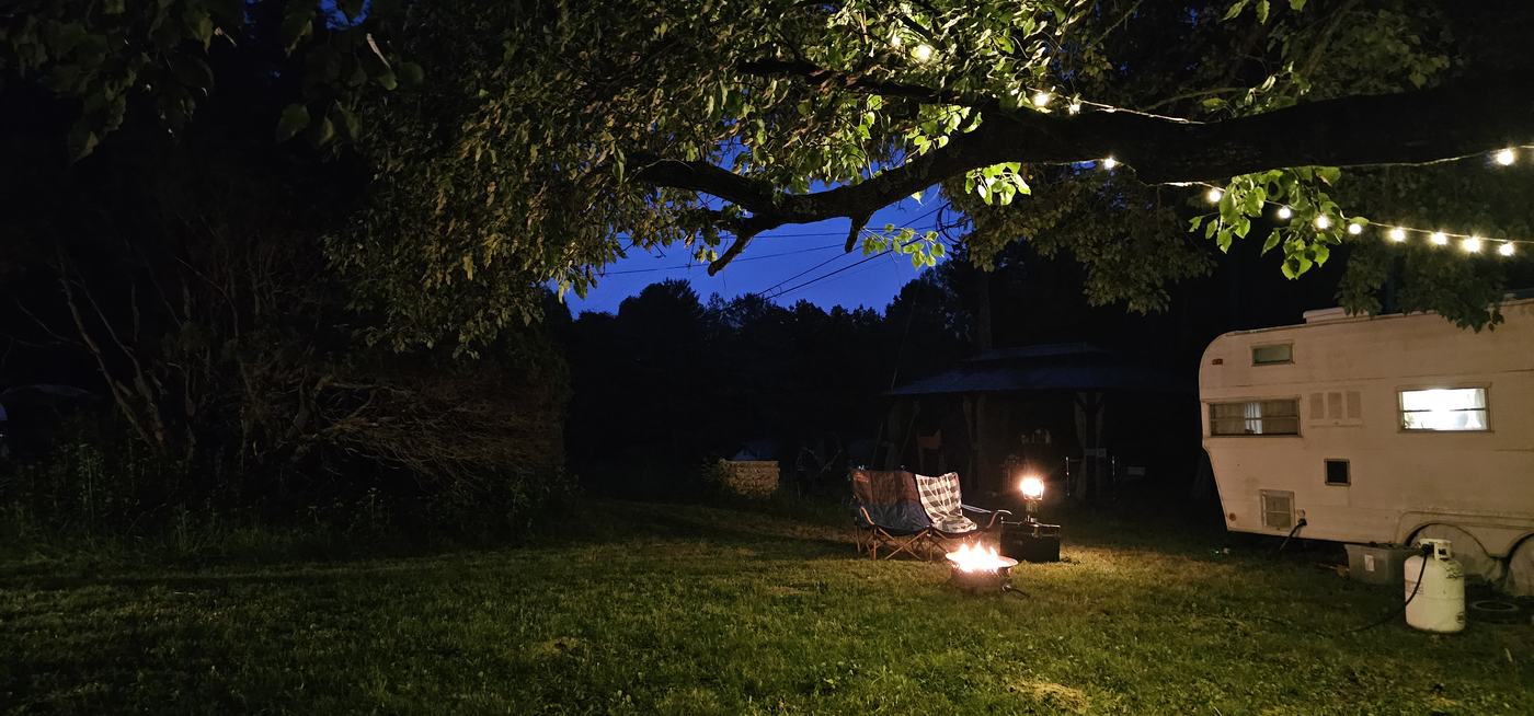 The ScandiFAN camper at dusk with a glowing window, a small fire, and string lights in the tree above